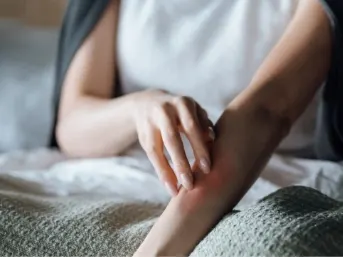 Stock photograph of a model: a woman sitting on a bed rubs a red, irritated patch on her forearm