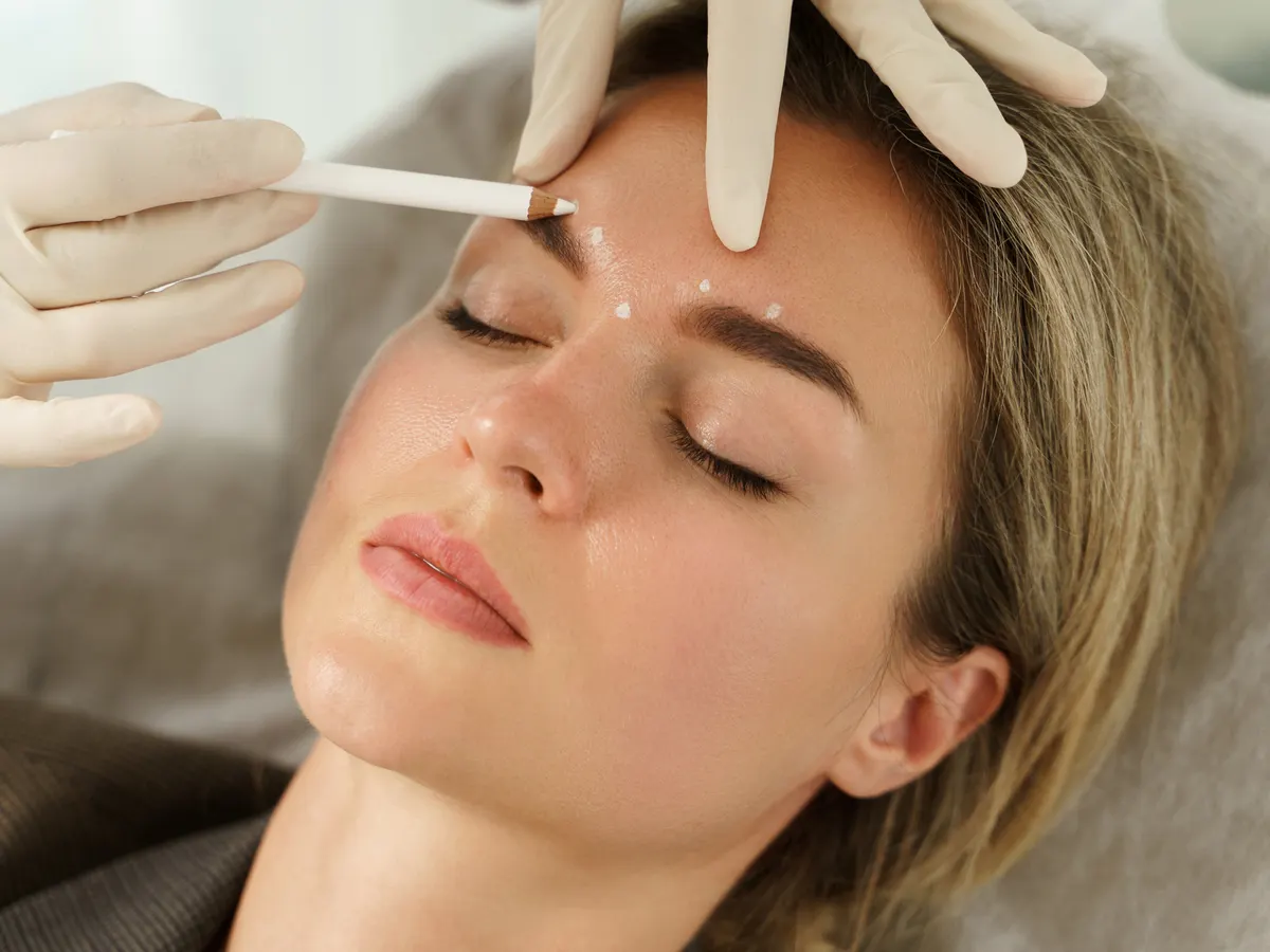 Stock photograph of a model: a woman lies back with her eyes closed while a gloved hand marks points on her forehead