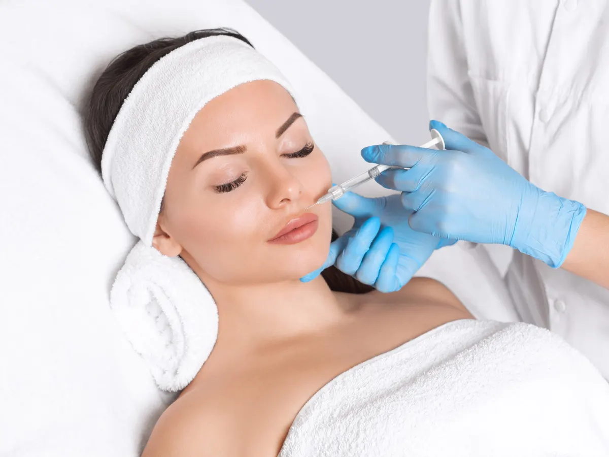 Stock photograph of models: a woman in a headband and towel lies back as a gloved clinician injects near her lip