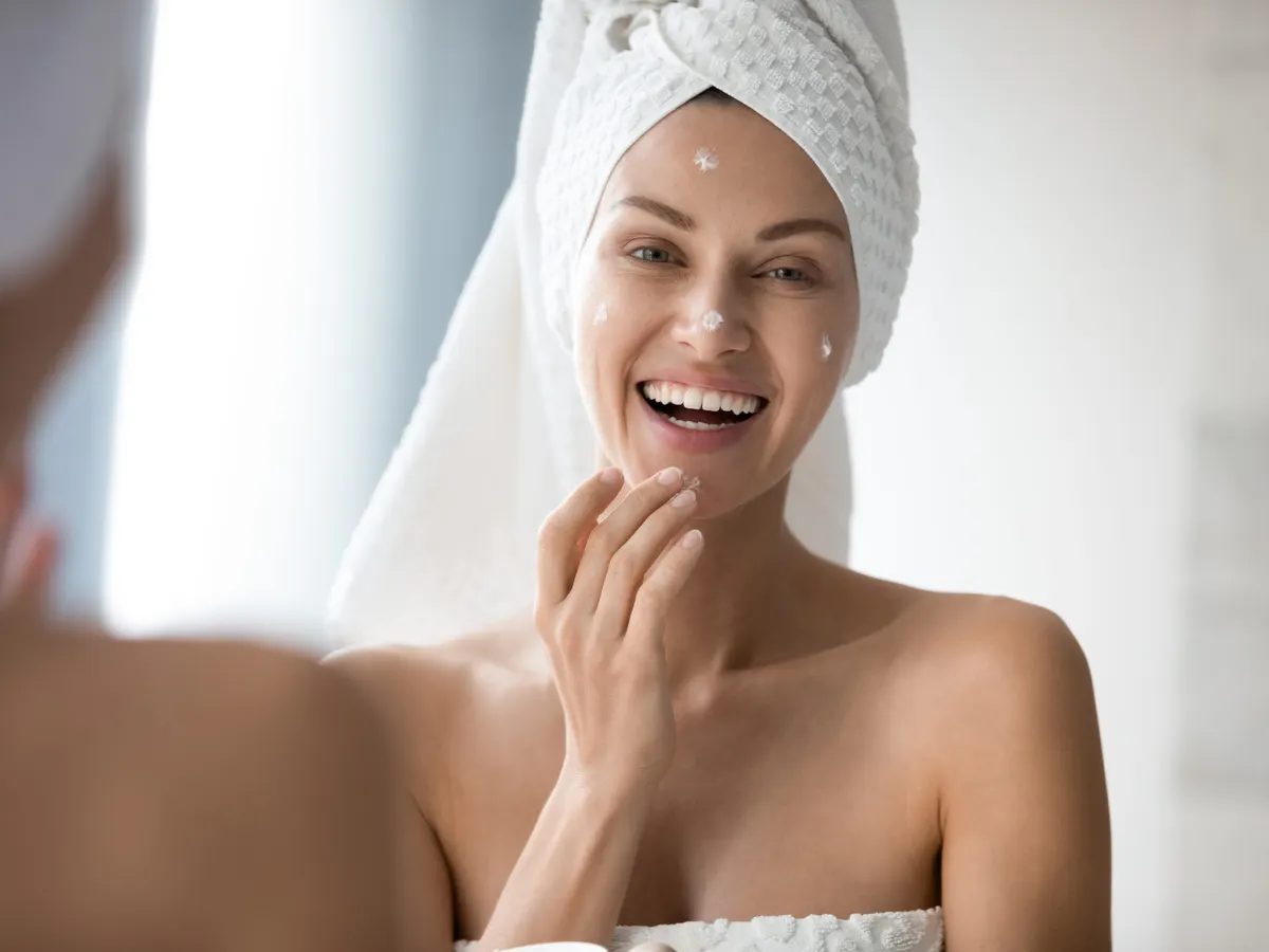 Woman in towel smiling while looking in mirror putting on moisturizer after microneedling in Jacksonville