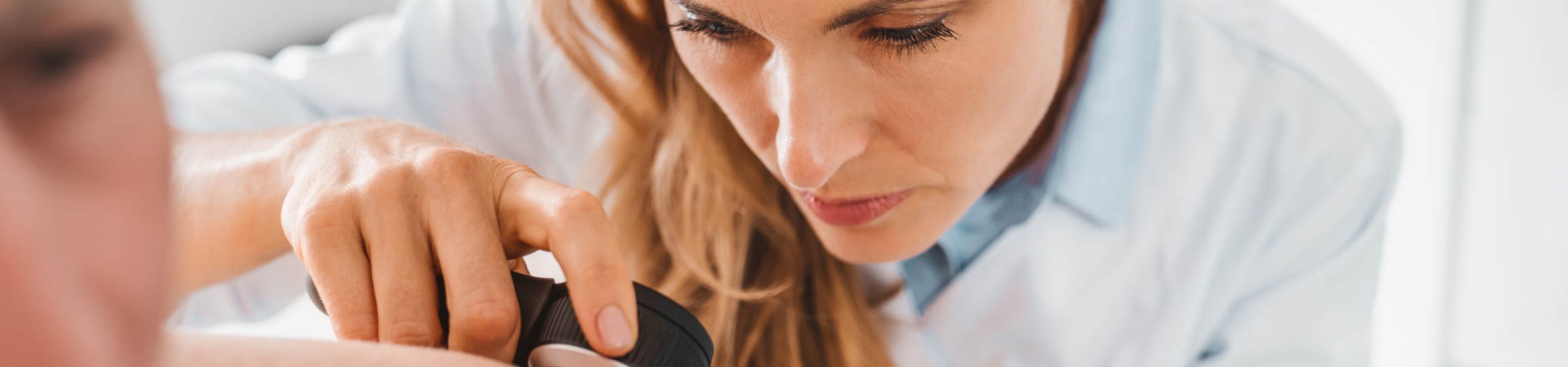 Stock photograph of models: a clinician holds a dermatoscope against a patient's skin and looks closely at it
