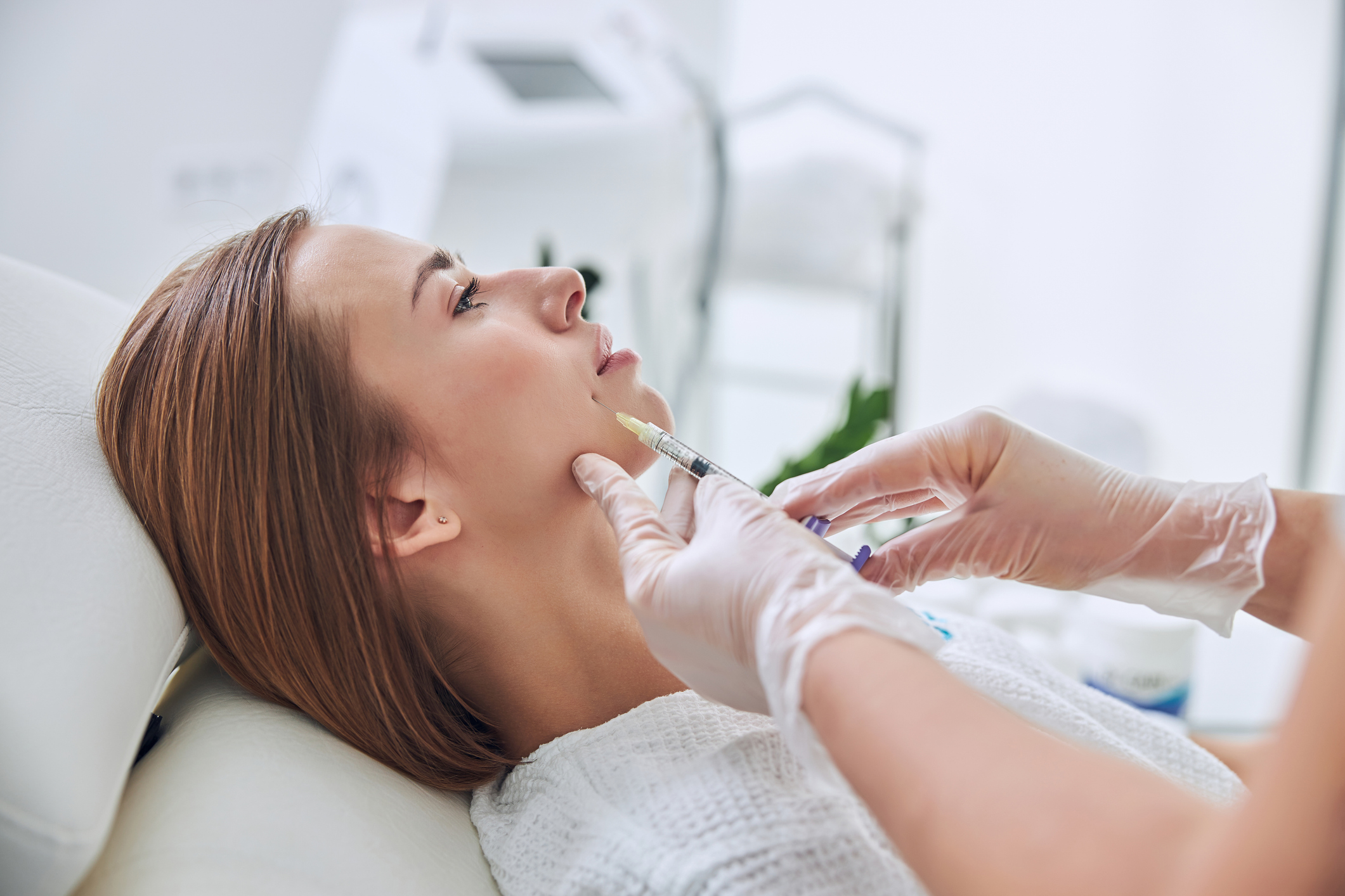 Stock photograph of models: a woman lies back while a gloved clinician injects filler near her chin