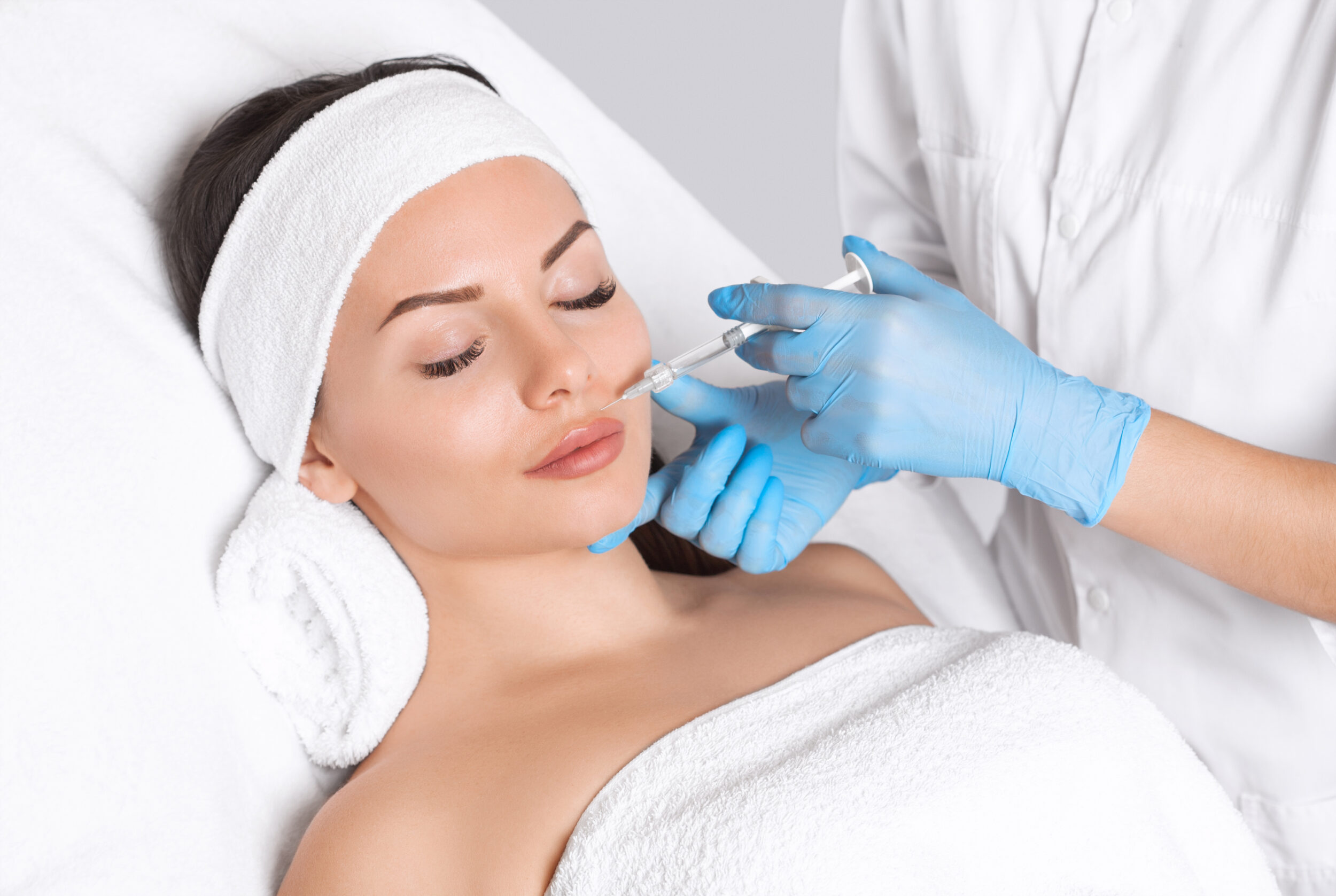 Stock photograph of models: a woman in a headband and towel lies back as a gloved clinician injects near her lip